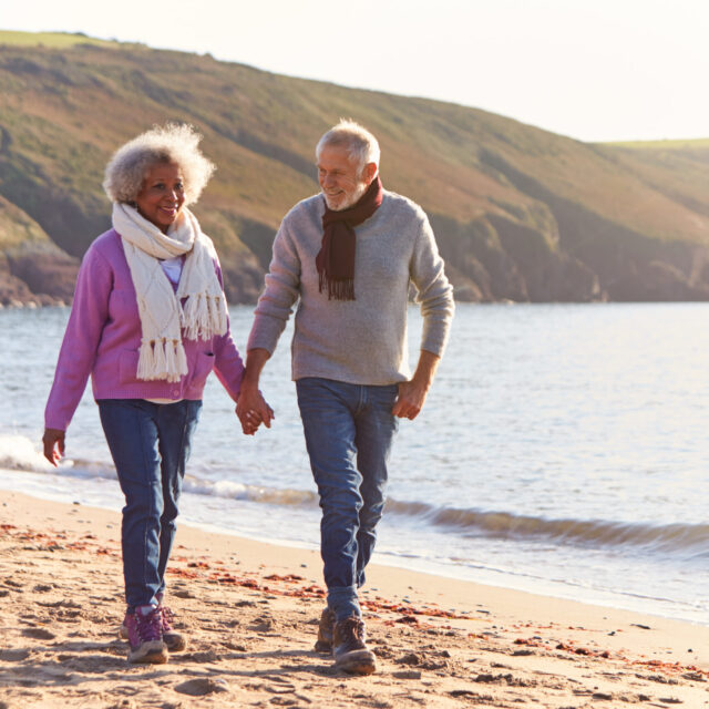 Loving Retired Couple Holding Hands As They Walk Along Shoreline On Winter Beach Vacation