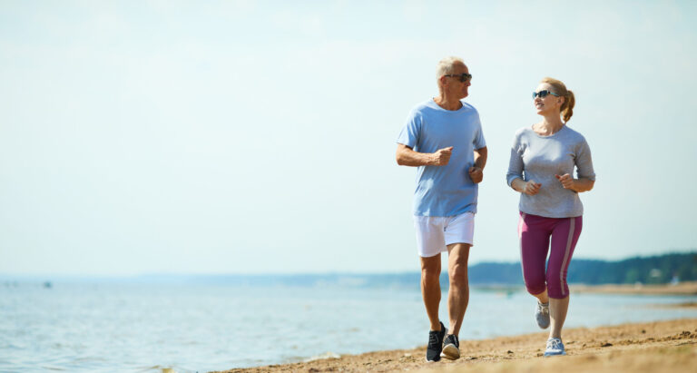 Active senior man and woman running down sandy beach with waterside on background